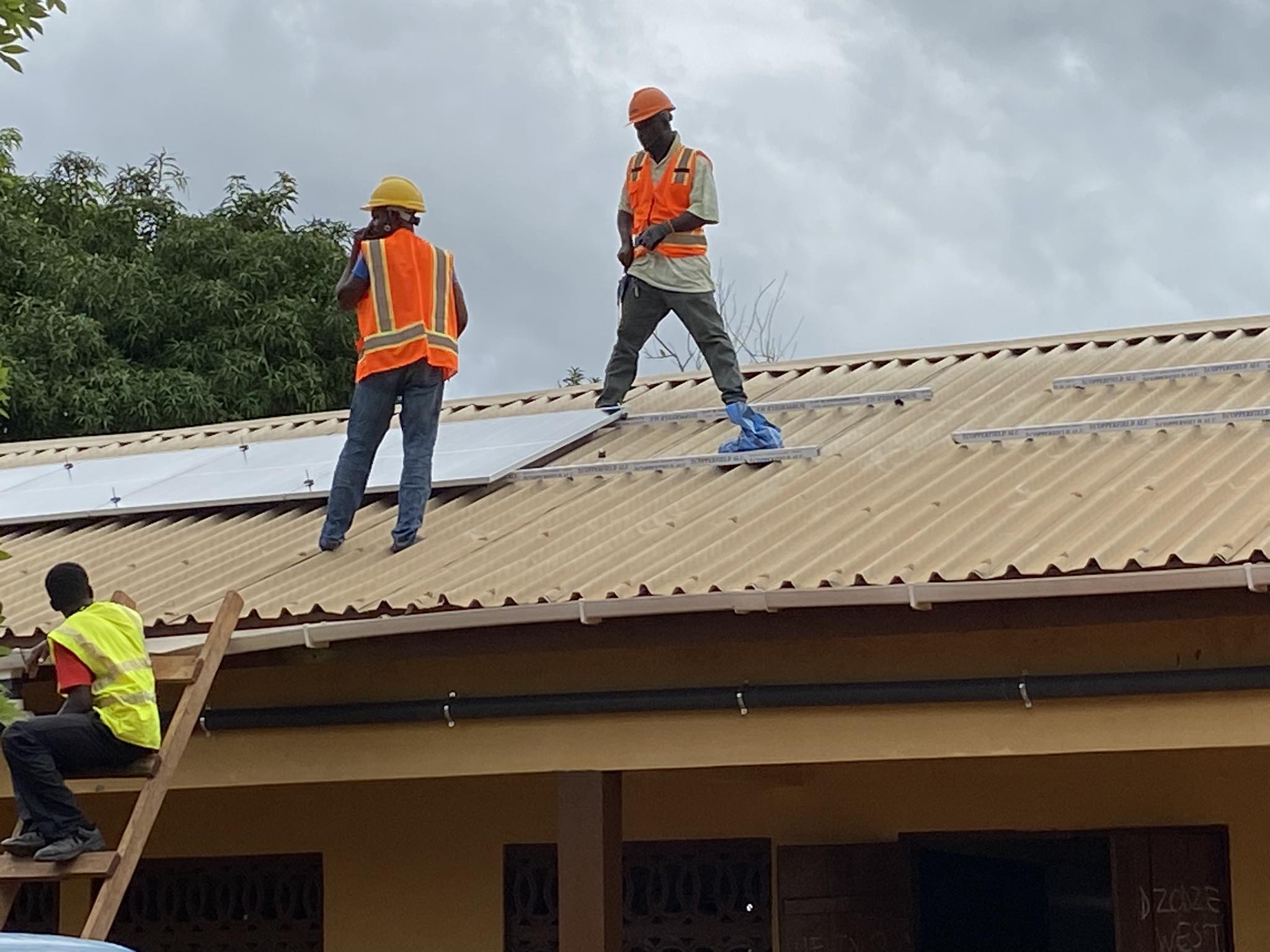 the roof of the classrooms has 2 men installing solar panels while a third man stands at the top of the ladder