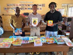 Abraham stands with 2 staff members behind a table covered with literacy reading sets.